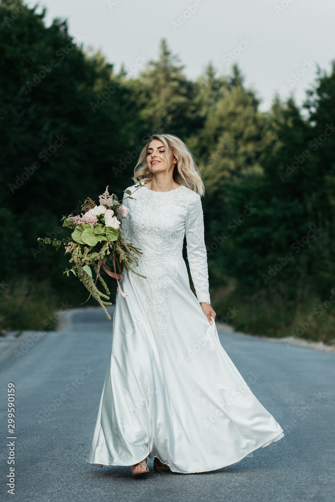 Beautiful elegant bride in lace wedding dress with long full skirt and long sleeves. She is holding a big bouquet of flowers. Outdoors, on the road.