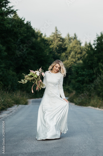 Beautiful elegant bride in lace wedding dress with long full skirt and long sleeves. She is holding a big bouquet of flowers. Outdoors, on the road. © Melika