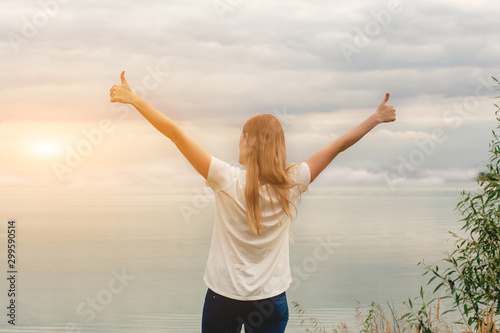 Beautiful girl on the lake. The view from the back. Hands up. Thumb up. Cloudy sky, sunset