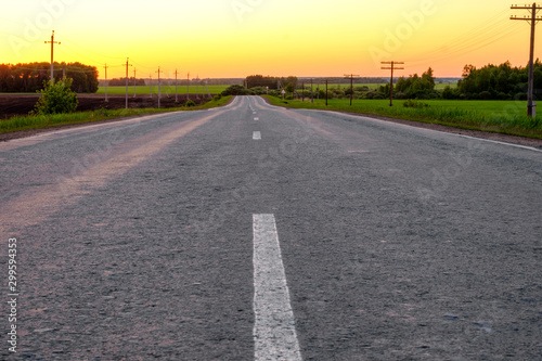 asphalt road stretching into the distance with markings on the edge of the road on both sides grass and electric poles the sky is red sunset