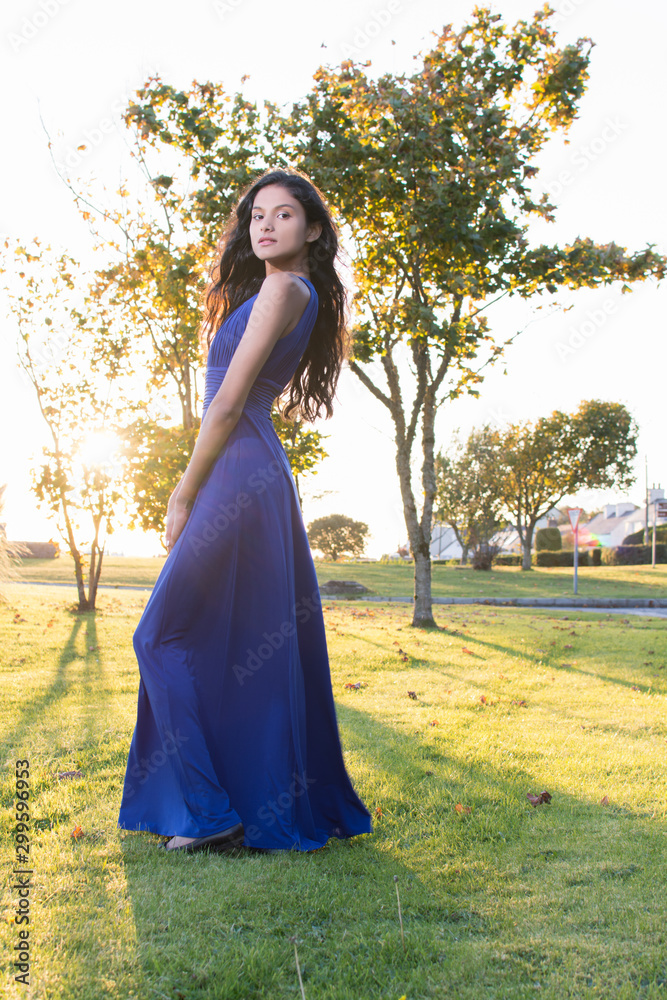 Beautiful model looking at camera in Sligo in Ireland. Full body. Mixed race Portuguese Indian female model posing. Blue dress. Standing on grass. Autumnal tree in the background. Lens flare effect
