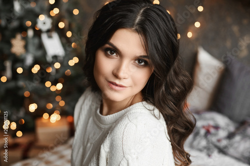 Close up portrait of a young beautiful smiling woman posing in an interior with festive Christmas lights on the background. Brunette model girl with trendy makeup and smooth skin looking in the camera
