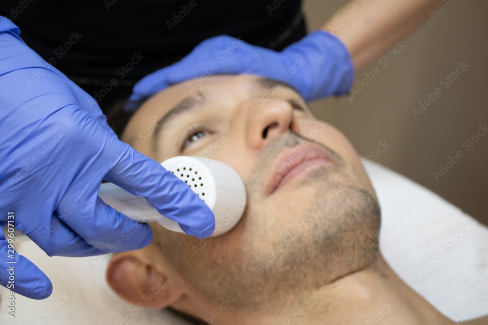 Young man laying on a facial therapy couch covered with the white towel with opened eyes. Facial therapist hands holding the facial machine above man face in a blue sterile gloves Young man laying on a facial therapy couch covered with the white towel with opened eyes. Facial therapist hands holding the facial machine above man face in a blue sterile gloves