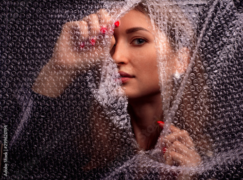 Abstract mysterious portrait girl hidden behind breaking bubble wrap in black background and looking through the gap plastic