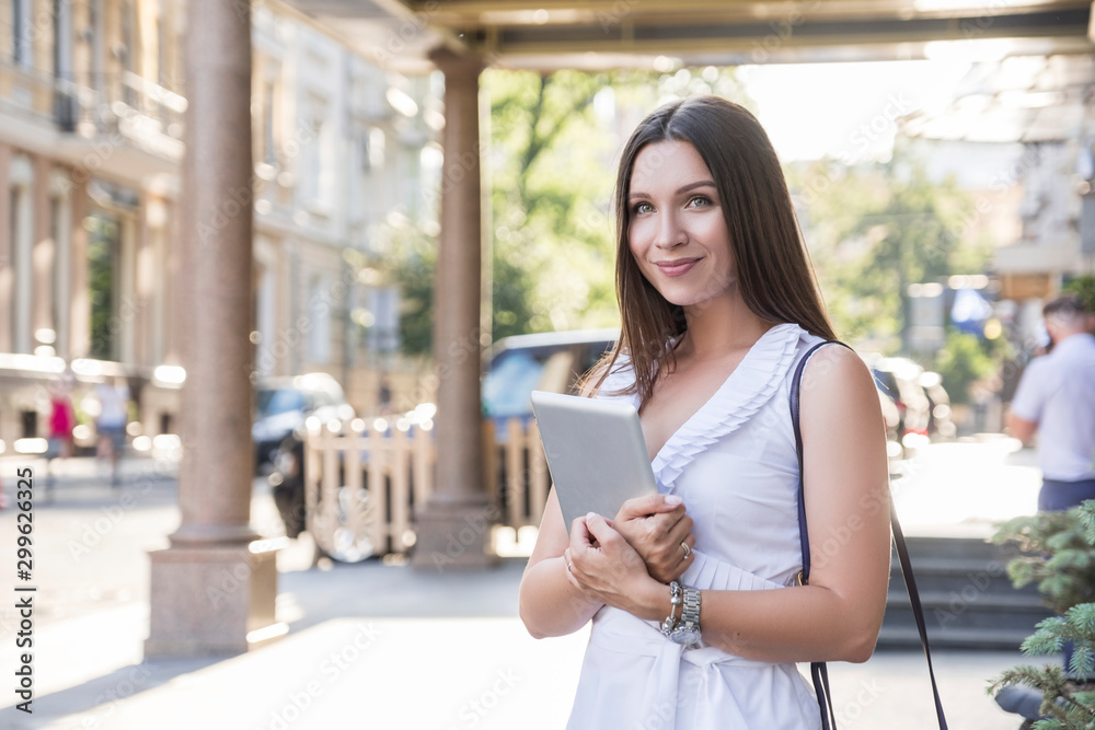 Young caucasian stylish woman in casual outfit on a european city street. She works on her tablet alone outdoors.