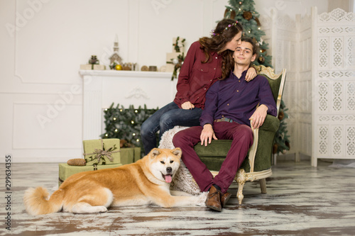 Young happy couple cuddling adorable akita inu dog while sit on stylish retro armchair for Christmas holidays at home. Christmas tree and fireplace background. Cozy home decorations © Kate