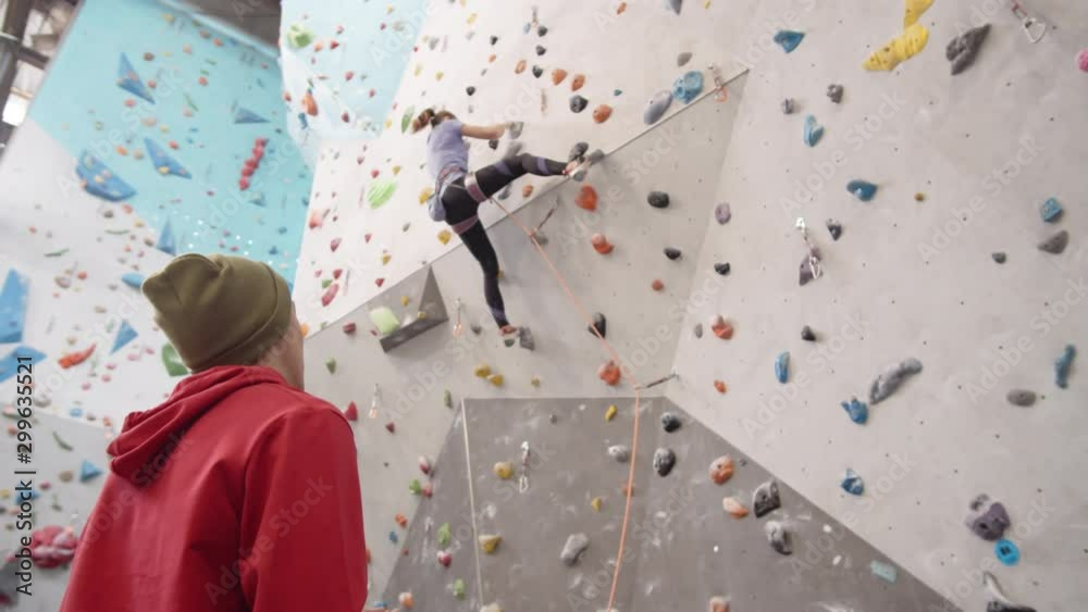 Panning of young man wearing red hoody and hat standing at climbing wall with rope in his hands and watching young Caucasian woman climbing artificial rock Panning of young man wearing red hoody and hat standing at climbing wall with rope in his hands and watching young Caucasian woman climbing artificial rock