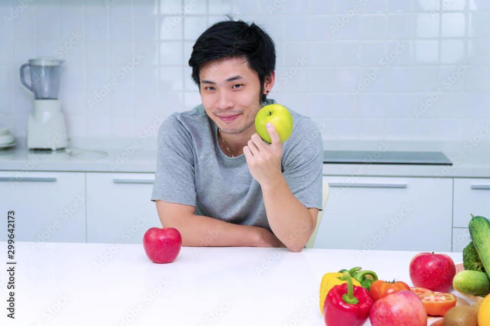 Healthy diet concept. Attractive smiling young man holding green fresh apple breakfast in his kitchen looking camera. Handsome male with vegetable and fruit on table at home. Healthy diet concept. Attractive smiling young man holding green fresh apple breakfast in his kitchen looking camera. Handsome male with vegetable and fruit on table at home.