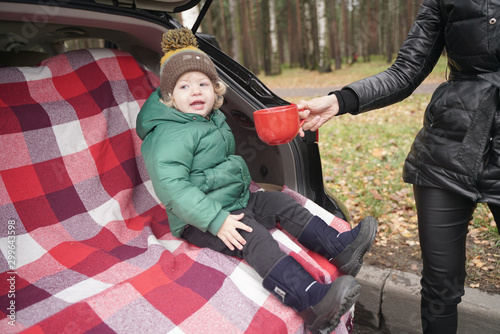 cute little kid boy sits in the trunk of a car on a red plaid and enjoys an autumn picnic alone