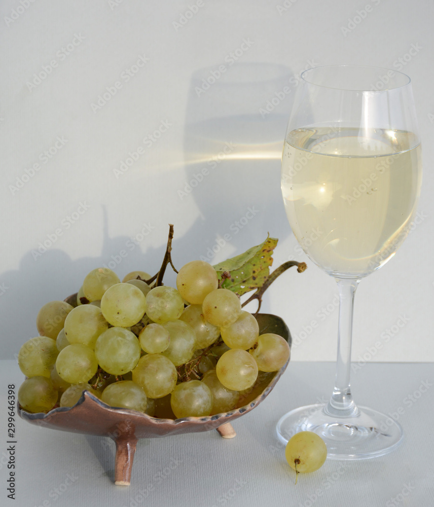 A glass of white wine with reflection on a white wall, a vase with grapes stands nearby A glass of white wine with reflection on a white wall, a vase with grapes stands nearby