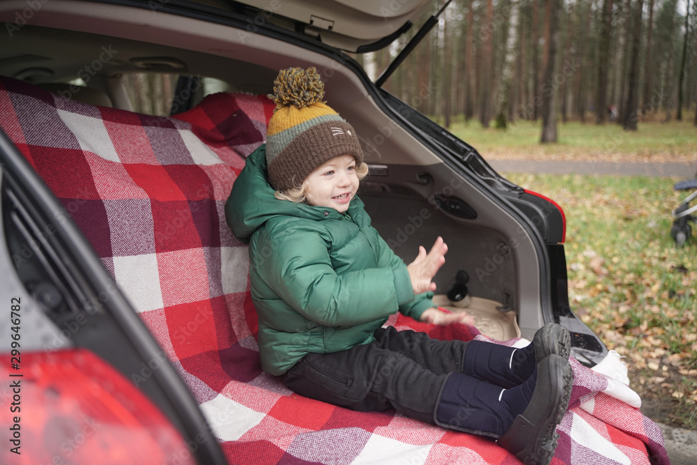 cute little kid boy sits in the trunk of a car on a red plaid and enjoys an autumn picnic alone
