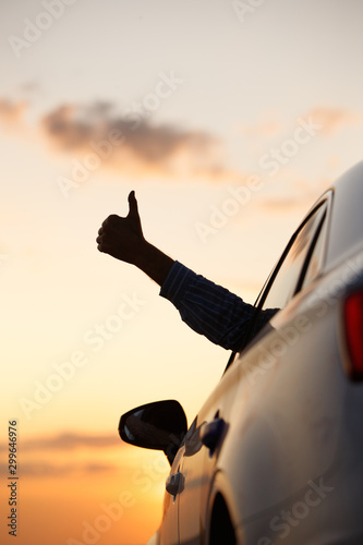 Man showing thumbs up/making Like / Ok sign with hand from car window with sunset sky, relaxing, enjoying road trip and feeling the air and freedom. Toward adventure, vacation 