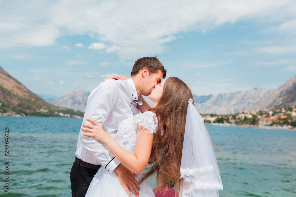 Wedding couple, newlyweds kiss on  background of sea and mountains. Groom and bride in white wedding dress with veil. Brunette man and brown-haired woman kissing outdoor