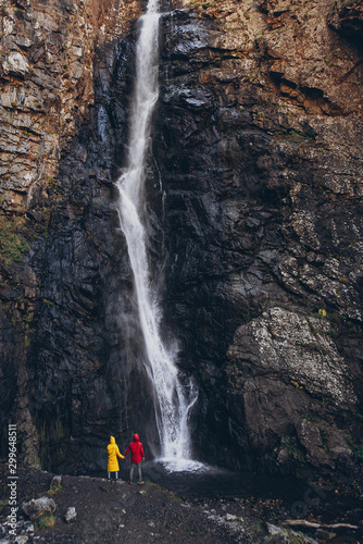 stylish tourist couple, man and woman dressed in bright raincoats of red and yellow are standing holding hands in front of a large waterfall