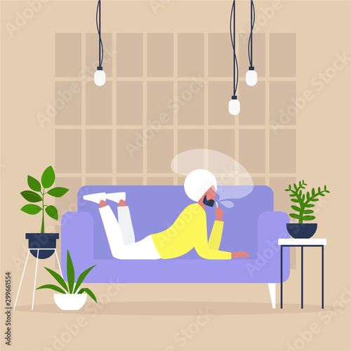 Young indian male character smoking a vape or a joint of cannabis on a sofa in a cozy interior with houseplants, modern millennial lifestyle