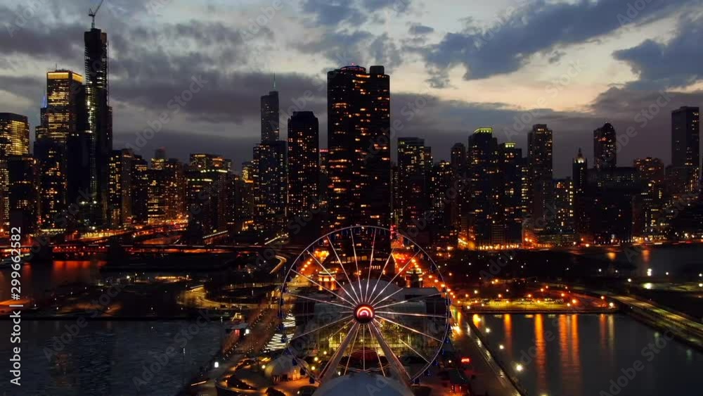 Aerial: Drone reversing over illuminated Ferris Wheel Navy Pier Chicago against modern skyscrapers, city against sky at night