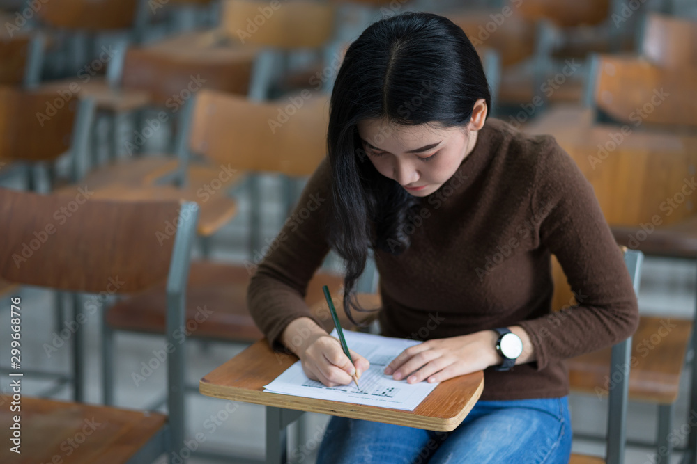 Young female university student concentrate doing examination in classroom. Girl student writes the exercise of the examinations. University classroom. Classroom chairs. University student. Young female university student concentrate doing examination in classroom. Girl student writes the exercise of the examinations. University classroom. Classroom chairs. University student.
