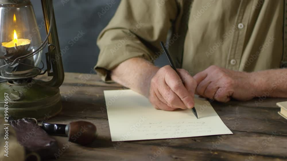 Handheld tilt up of romantic old-fashioned senior man in straw hat writing poem, then looking up and thinking. Smoking pipe lying beside him and lit oil lamp standing on wooden table Handheld tilt up of romantic old-fashioned senior man in straw hat writing poem, then looking up and thinking. Smoking pipe lying beside him and lit oil lamp standing on wooden table