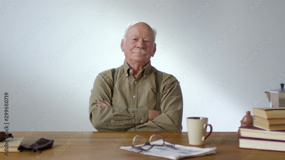 Dolly-in portrait shot of senior man with grey hair and mustache sitting at table with his arms crossed and looking at camera Dolly-in portrait shot of senior man with grey hair and mustache sitting at table with his arms crossed and looking at camera