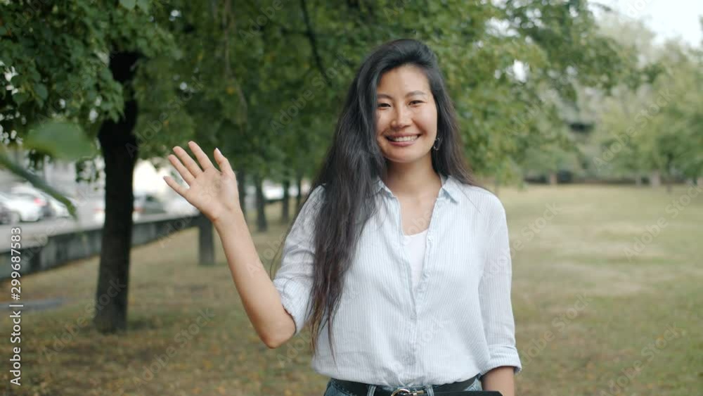 Slow motion portrait of young Asian girl waving hand smiling looking at camera outdoors in urban park. Millennials, positive emotions and happy youth concept. Slow motion portrait of young Asian girl waving hand smiling looking at camera outdoors in urban park. Millennials, positive emotions and happy youth concept.