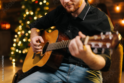 Happy young man is playing guitar. Guy is looking happily and carefree. Male in festive hat alone celebrating Christmas or new year. Christmas tree with garland in background.