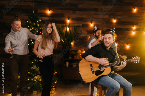 Happy young people are dancing and singing at the Christmas party in cozy house. Happy young man plays guitar. Christmas tree with garland and wall with festive illumination in background