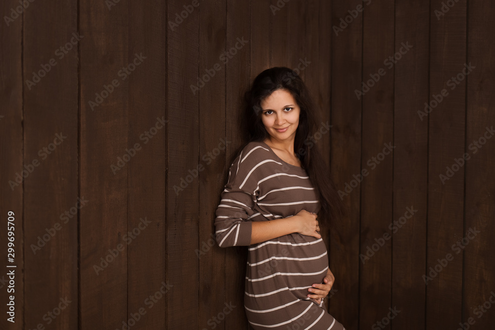 Fototapeta premium Pregnant woman in brown stripe dress holds hands on belly on a dark brown background. Pregnancy, maternity, preparation and expectation concept. Beautiful tender mood photo of pregnancy