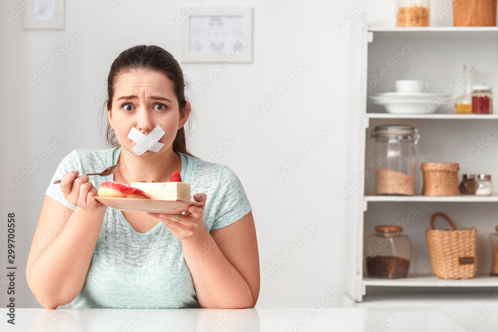 Junk Food. Chubby girl taped with patch mouth sitting at kitchen table with desserts looking camera unhappy - obrazy, fototapety, plakaty 
