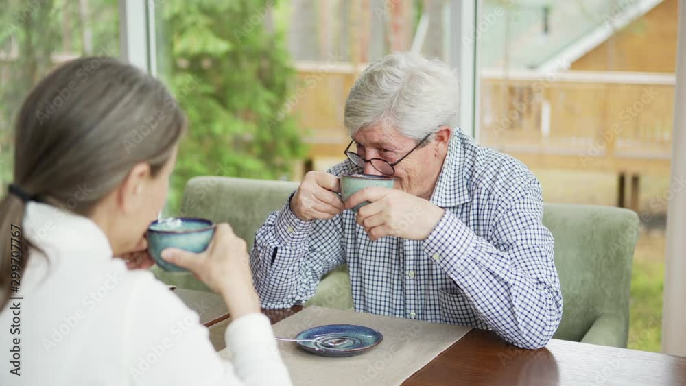 Over the shoulder view of senior man in eyeglasses talking with wife or female friend over tea or coffee sitting at table in cafe Over the shoulder view of senior man in eyeglasses talking with wife or female friend over tea or coffee sitting at table in cafe