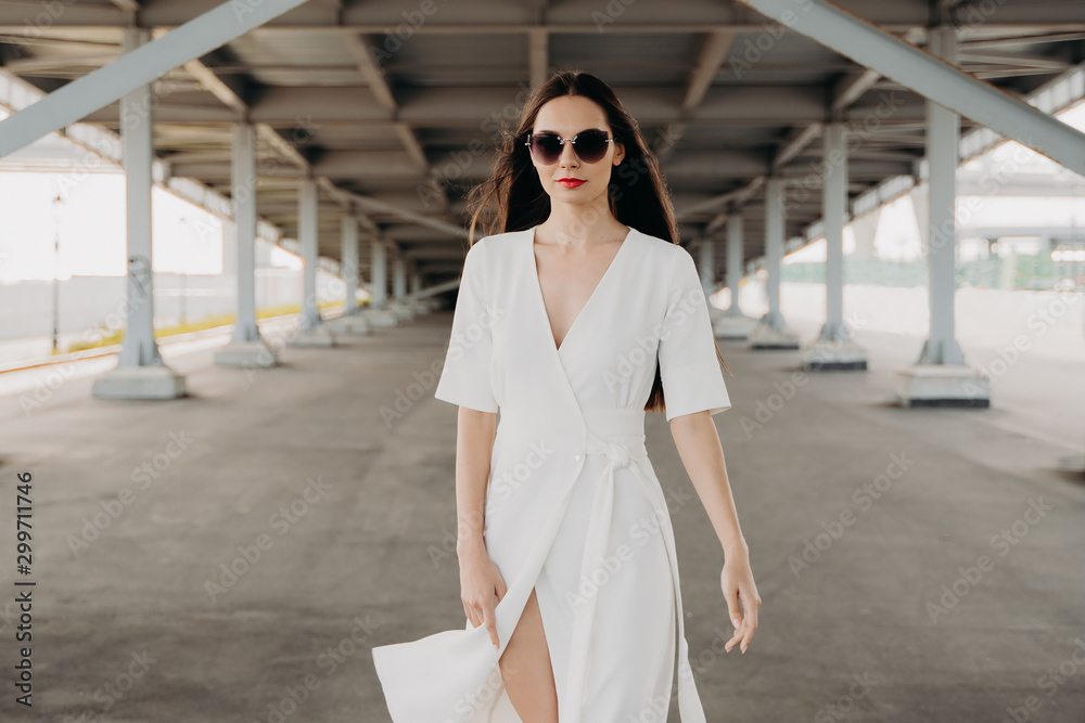 Fototapeta premium young woman in the city. Portrait of a young woman in sunglasses. The girl in the white dress. Walk around the city. Long-haired brunette. Female portrait. Beauty and fashion. Gray background. Bridges