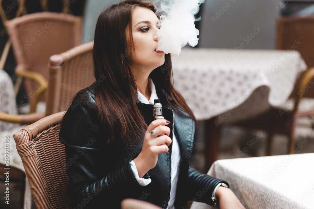 Pretty lady holding an electronic cigarette in her hand while sitting at a cafe table. Woman relaxes smoking vape and blowing thick smoke from the mouth.