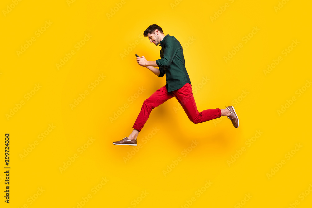 Full size profile side photo of positive cheerful brunet hair use his cellphone search find discounts jump run feel content wear green shirt red pants trousers isolated over yellow color background