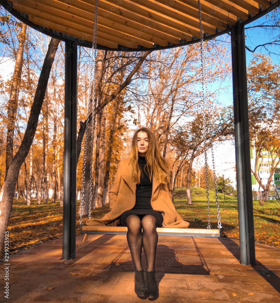 Girl swinging on a wooden swing hanging on chains in the autumn park. A young woman sits on a swing in a city park. Girl swinging on a wooden swing hanging on chains in the autumn park. A young woman sits on a swing in a city park.