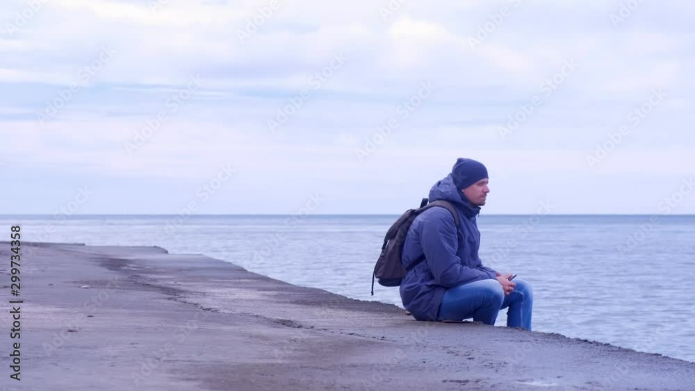 Young man traveller with backpack and smartphone in hands wears in jacket and hat at sea. Man tourist comes to old sea waterfront sits and looks at sea in winter. Overcast day at se.
