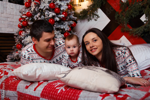 family in pajamas sits on a bed near the Christmas tree mom, dad and toddler, idyll, Christmas celebration. family happiness.
