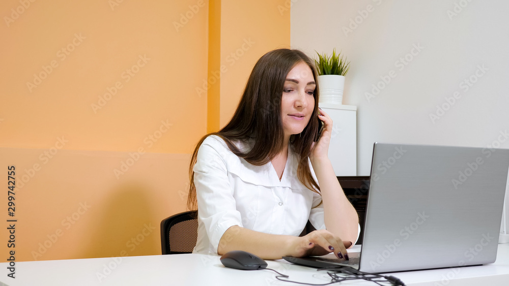 smiling lady receptionist in white coat talks on smartphone explaining to customer price list at laptop in beauty salon, copyspace smiling lady receptionist in white coat talks on smartphone explaining to customer price list at laptop in beauty salon, copyspace