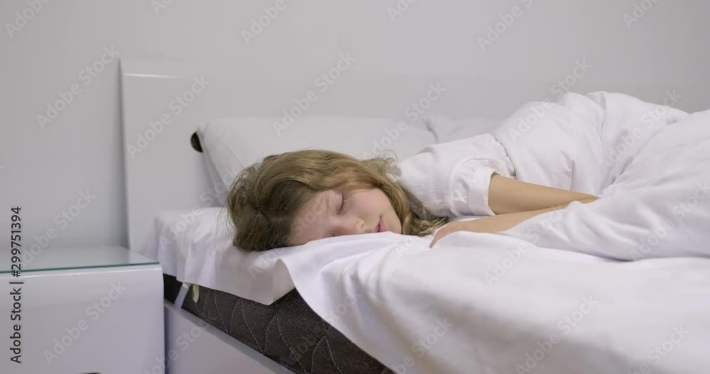 Girl sleeping in white bed. Waking up, touching sleepy eyes with hands, child wants to sleep Girl sleeping in white bed. Waking up, touching sleepy eyes with hands, child wants to sleep