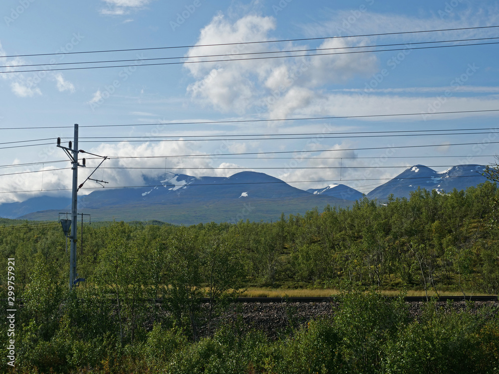 railroad in Abisko national park with birch forest and snow capped mountains, summer blue sky clouds