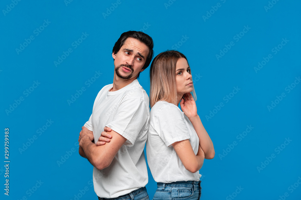 Fototapeta premium Upset couple of a young blond woman and brunet bearded man with mustaches in white t-shirts and blue jeans posing isolated over blue background.