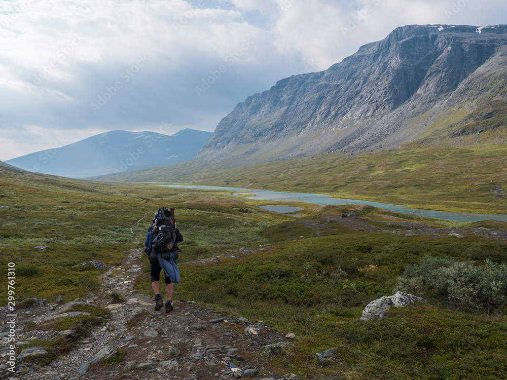 Man hiker with heavy backpack in wild Lapland nature with blue glacial river, birch tree bushes, snow capped mountains and dramatic clouds. Northern Sweden summer at Kungsleden hiking trail.