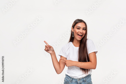 Pretty african-american teenager in a white t-shirt and blue jeans pointing at the empty space in a left side of a frame standing isolated over white background.