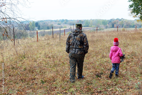 A father in camouflage walks with his young daughter in a red jacket and a cap with a bubo on dry grass in the countryside. Beautiful view of the hills with autumn forest.