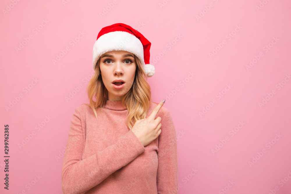 Portrait of surprised girl in christmas hat and pink sweater stands on pink background, points her finger to the empty space and looks with shocked face into the camera. Christmas sale. Portrait of surprised girl in christmas hat and pink sweater stands on pink background, points her finger to the empty space and looks with shocked face into the camera. Christmas sale.