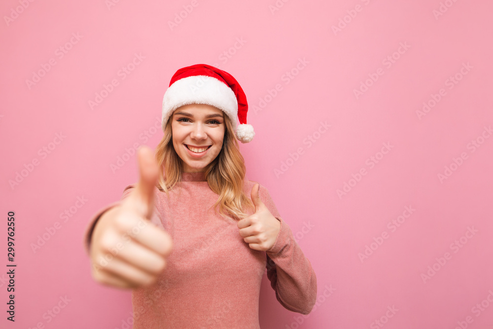 Happy blonde girl in santa hat and pink sweater stands on pink background and shows thumbs up, looks at camera and smiles. Smiling lady in santa hood shows gesture like. Isolated. X-mas