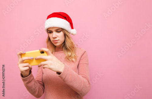 Attractive blonde girl plays mobile games on a smartphone with a serious face looks intently at the phone screen  wears a santa claus hat and a pink sweater.Girl gamer plays mobile games for Christmas