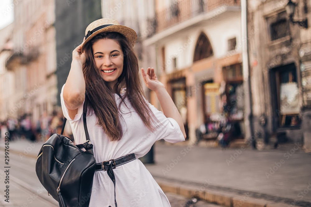 Fototapeta premium Young stylish woman walking on the old town street, travel with backpack, straw hat, wearing trendy outfit.