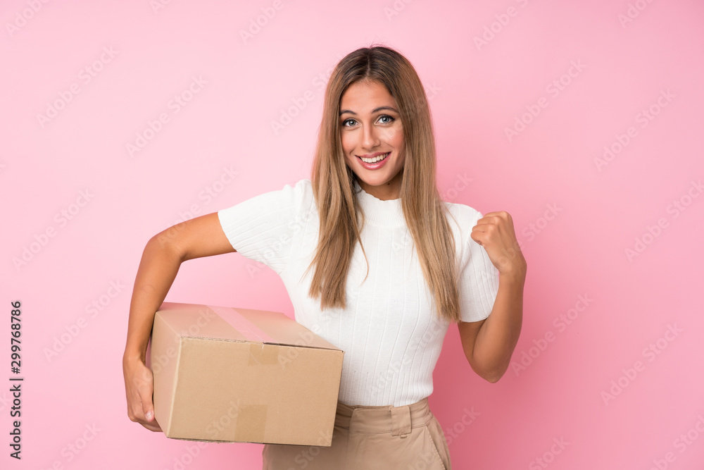 Young blonde woman over isolated pink background holding a box to move it to another site Young blonde woman over isolated pink background holding a box to move it to another site