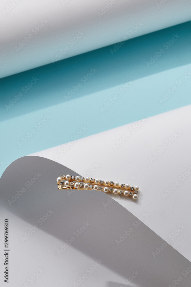 Object photo of a golden rectangular alligator hair pin, decorating with white beads and rhinestones, lying on a white half-cylinder. The photo was taken on a blue background.  