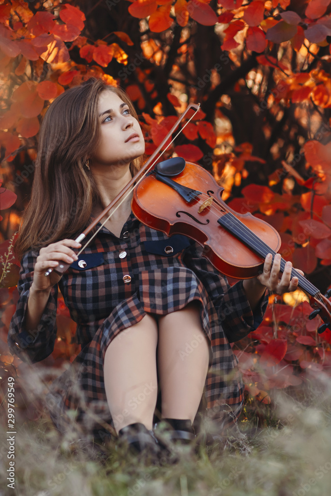 autumn portrait of beautiful woman sitting on the ground with a violin under chin on a background of red foliage, girl engaged in playing a musical instrument in nature, a concept of passion in art