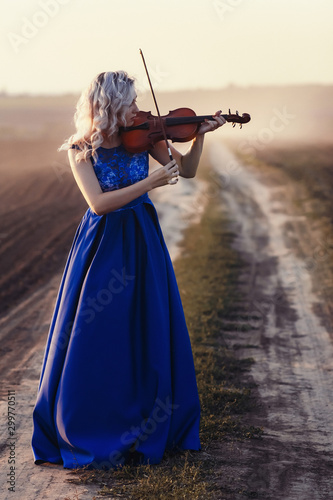 woman in long dress playing violin on background of field path with a club of dust, girl engaged in musical art, performance on nature, concept passion in music photo